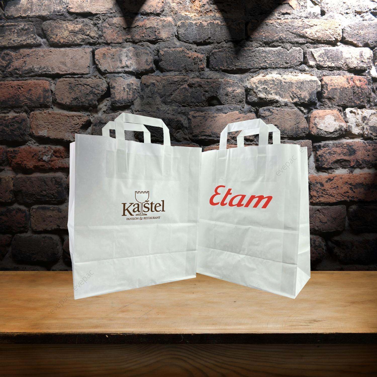 Sacs en papier kraft personnalisés blancs avec poignées plates posés sur une table en bois, devant un mur de briques.